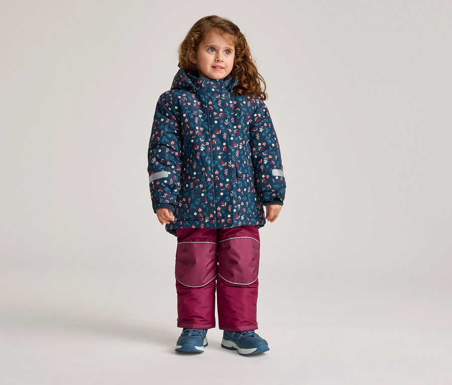 Une fillette aux cheveux bouclés se tient debout, portant une veste bleue à fleurs et un pantalon bordeaux.
