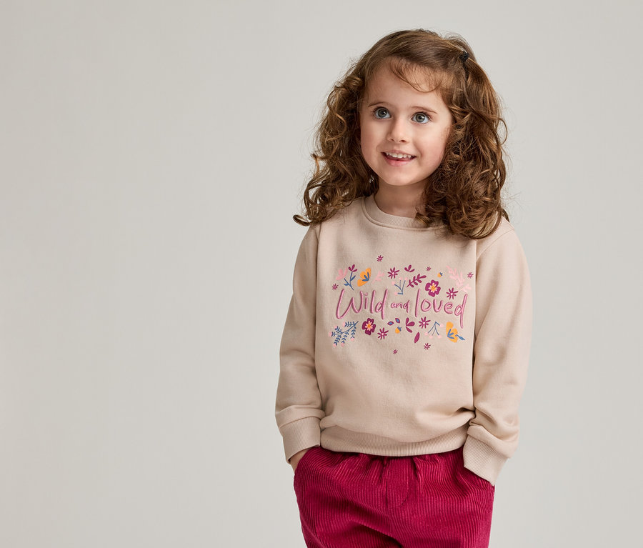 Une fillette aux cheveux bouclés porte un pull marron clair avec l'inscription « Wild and loved » et un pantalon rouge.
