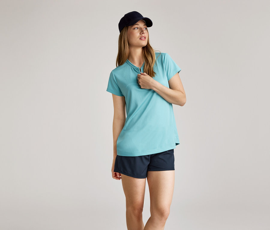 Femme portant une casquette bleu marine, un t-shirt bleu clair à fermeture éclair courte et un short bleu marine sur fond clair.