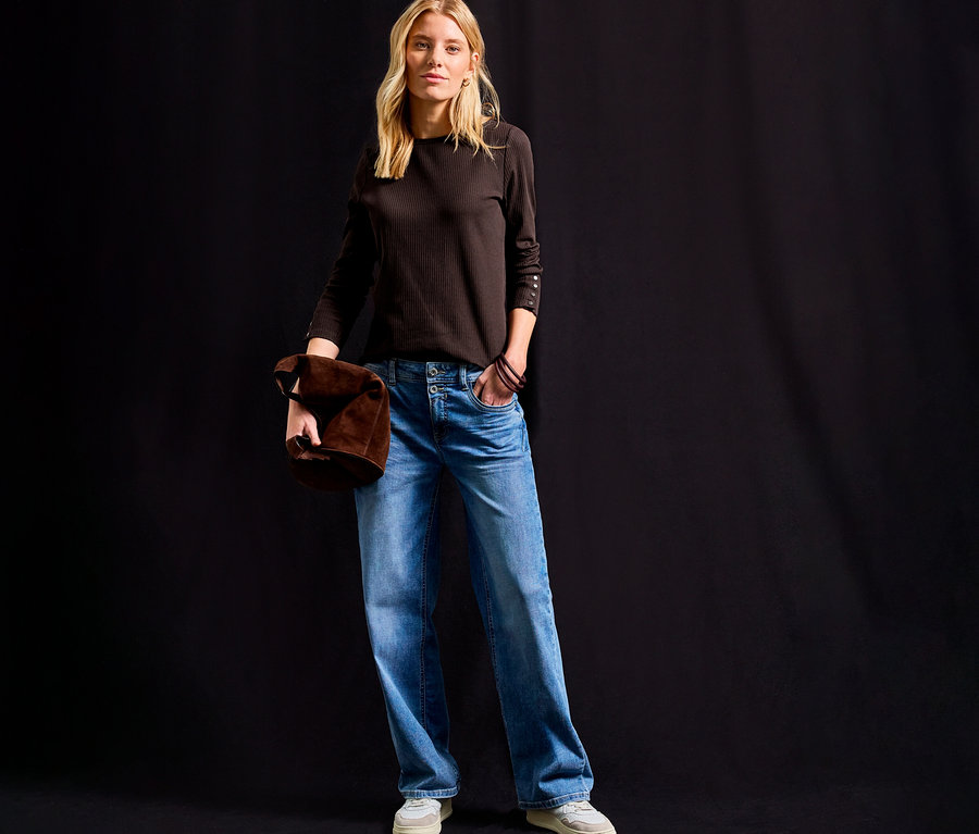 Femme debout tenant un sac marron, portant un maillot côtelé Street One marron, un jean et des chaussures claires.