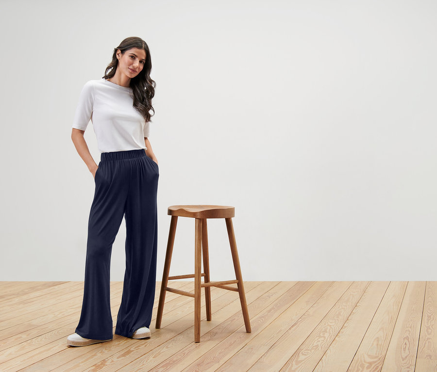 Femme debout à côté d'une chaise en bois, portant un t-shirt blanc et un pantalon large bleu marine.