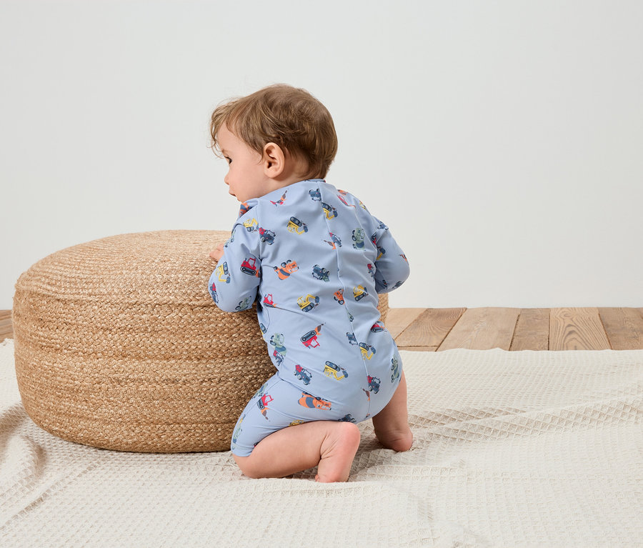 Un bébé en combinaison bleu clair à motifs de véhicules de chantier est agenouillé sur une couverture blanche à côté d'un pouf rond tressé.