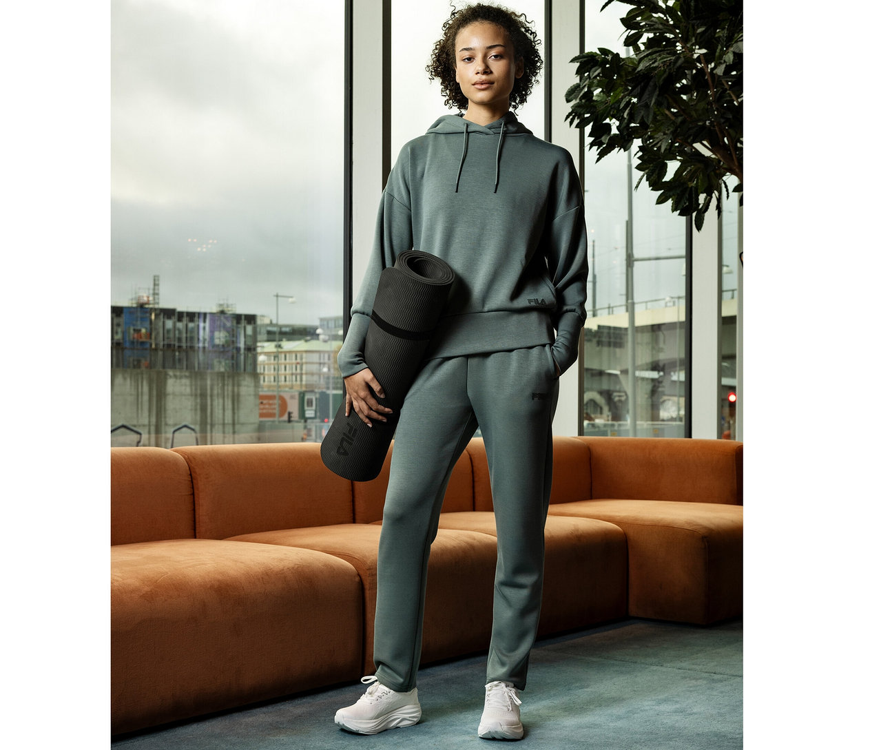 Femme en pantalon de jogging FILA vert sauge en mélange de modal et sweat à capuche, tenant un tapis de yoga noir, devant une fenêtre.