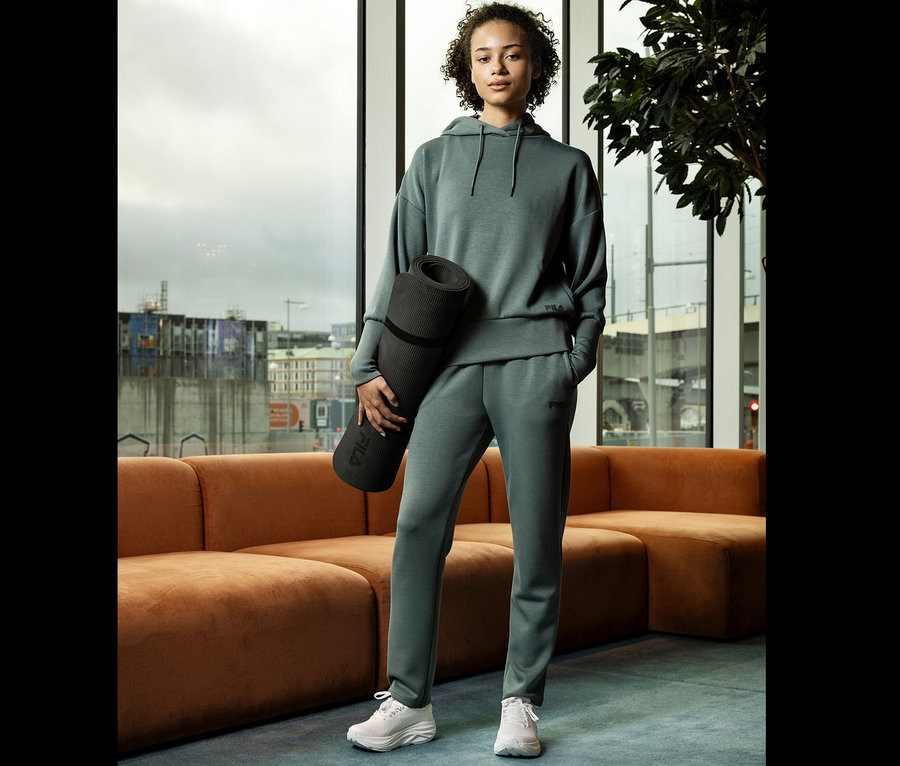 Femme en survêtement vert tenant un tapis de yoga devant une fenêtre.