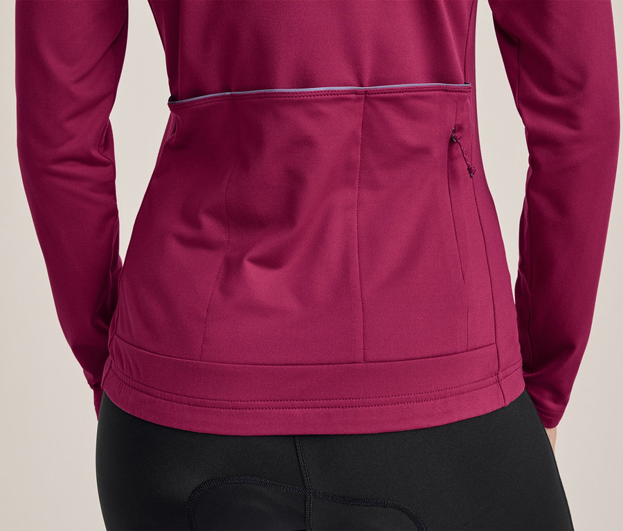 Personne de dos portant un maillot de cyclisme à manches longues magenta et un cuissard noir. Le maillot a trois poches arrière, dont une zippée, et une bande réfléchissante.