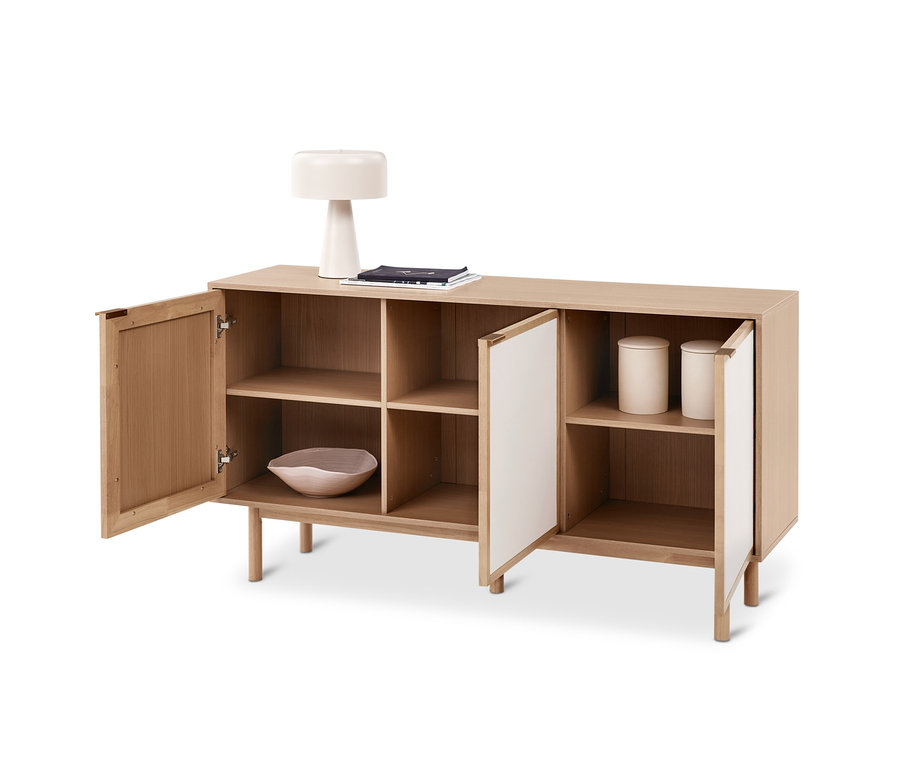 Un buffet en bois clair à quatre compartiments, les portes extérieures sont ouvertes. Une lampe blanche et des livres sont dessus, un bol et deux récipients sont à l'intérieur.