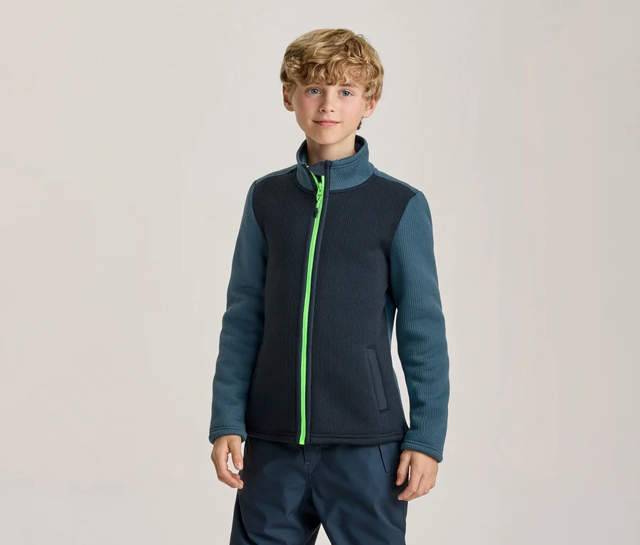 Jeune garçon debout, regardant la caméra. Il porte une veste bleu foncé avec des manches sarcelle et une fermeture éclair vert fluo, associée à un pantalon bleu foncé.