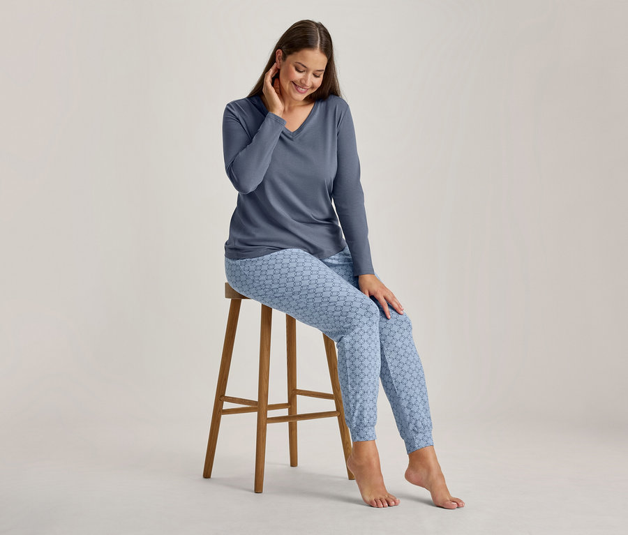 Une femme souriante est assise sur un tabouret en bois, vêtue d'un haut à manches longues et col en V et d'un pantalon de pyjama bleu clair à motifs. Elle regarde vers le bas et touche son cou.