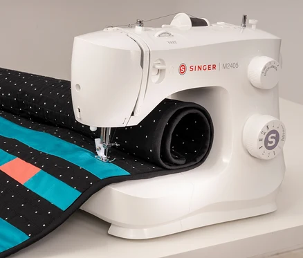 Une machine à coudre blanche Singer M2405 coud un tissu avec des rayures bleues et orange.