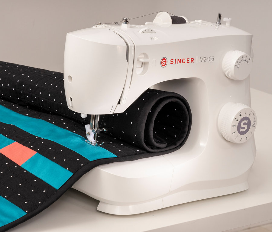 Une machine à coudre blanche Singer M2405 coud un tissu avec des rayures bleues et orange.