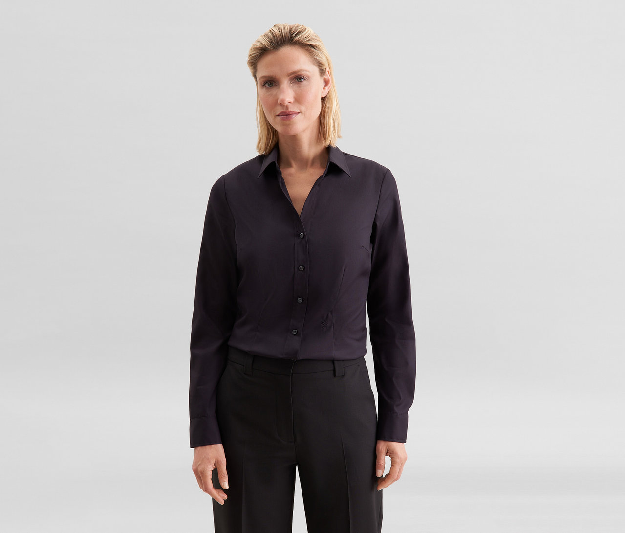 Femme blonde portant une chemise foncée à manches longues et un pantalon foncé, regardant l'appareil photo.
