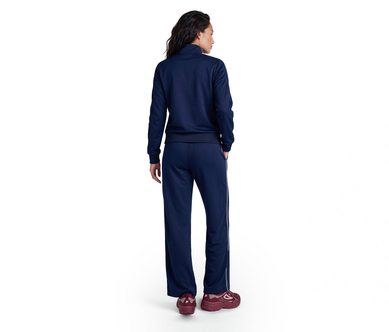 Une personne de dos, portant un survêtement bleu marine avec une veste zippée et un pantalon large à bande latérale blanche, ainsi que des baskets bordeaux.