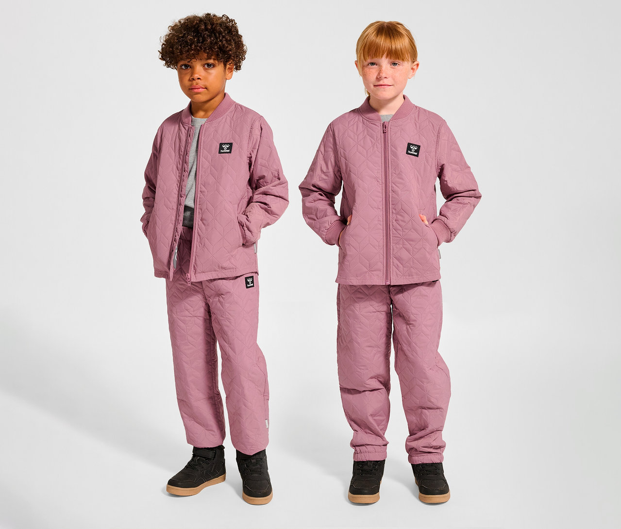 Deux enfants, un garçon et une fille, se tiennent côte à côte, vêtus d'ensembles thermiques hummel® HMLSOBI roses assortis, composés de vestes et de pantalons matelassés.