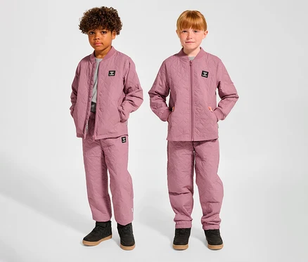 Deux enfants, un garçon et une fille, se tiennent côte à côte, vêtus d'ensembles thermiques hummel® HMLSOBI roses assortis, composés de vestes et de pantalons matelassés.