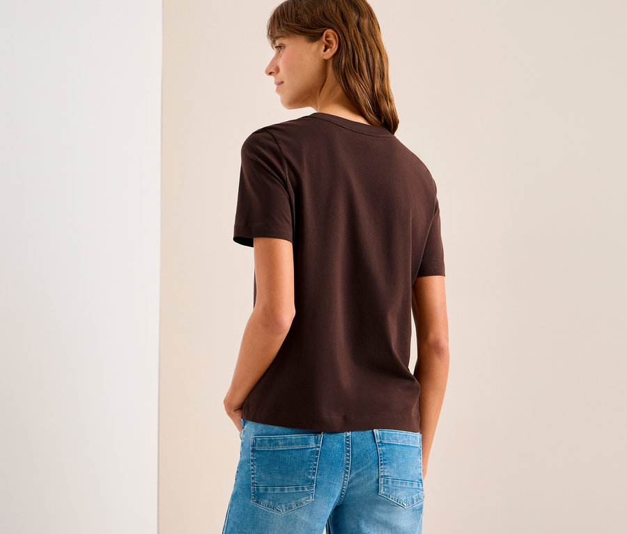 Femme portant un T-shirt à motif CECIL marron et un jean bleu, les mains dans les poches, regardant vers la gauche devant un mur blanc et beige.