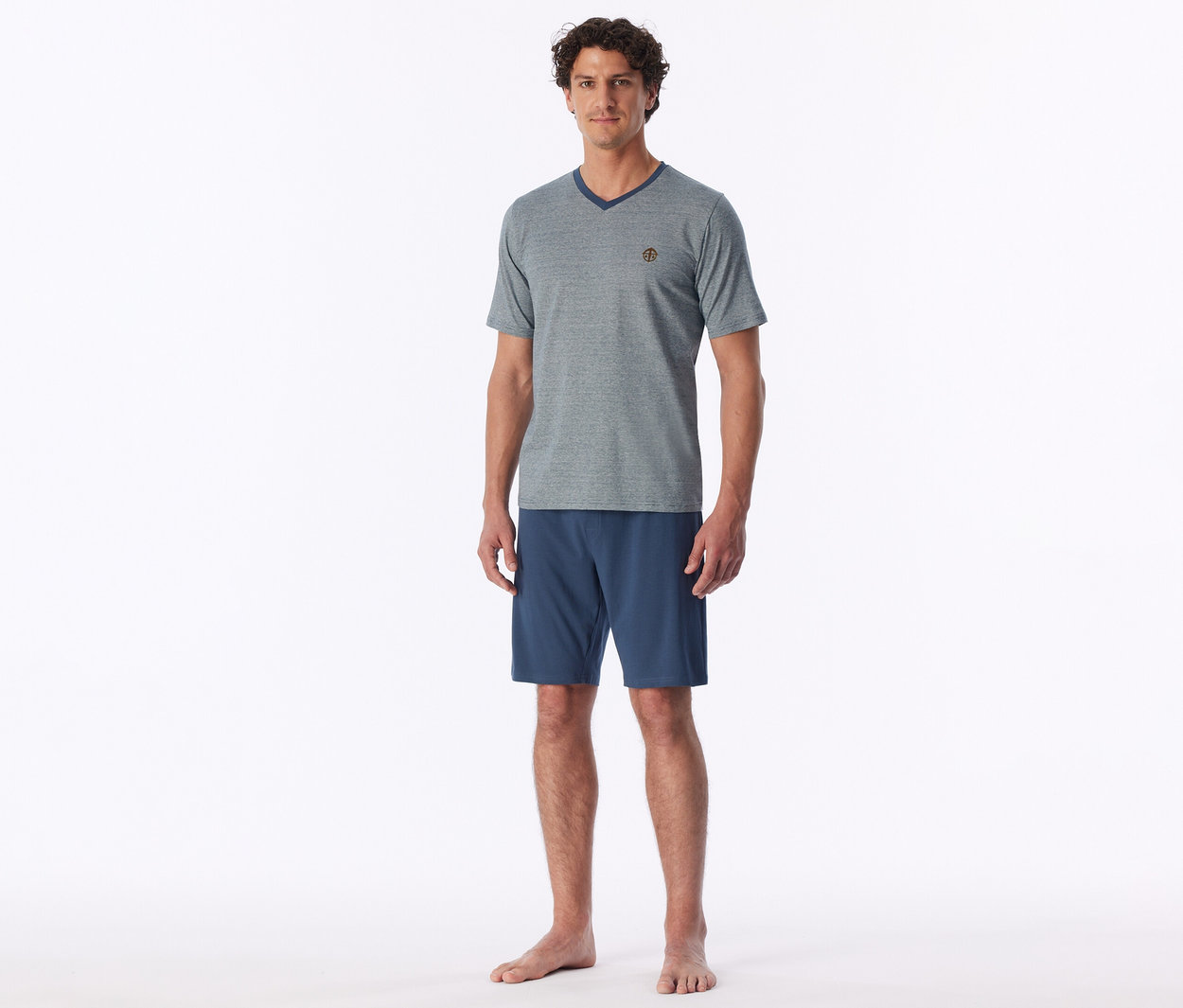 Un homme debout pieds nus, portant un t-shirt gris à col en V et un short bleu.