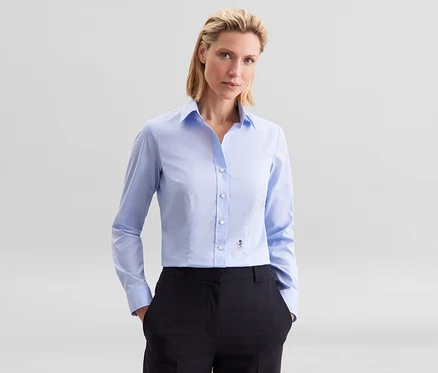 Une femme porte un chemisier Seidensticker Slim Fit non iron bleu clair et un pantalon noir, les mains dans les poches.