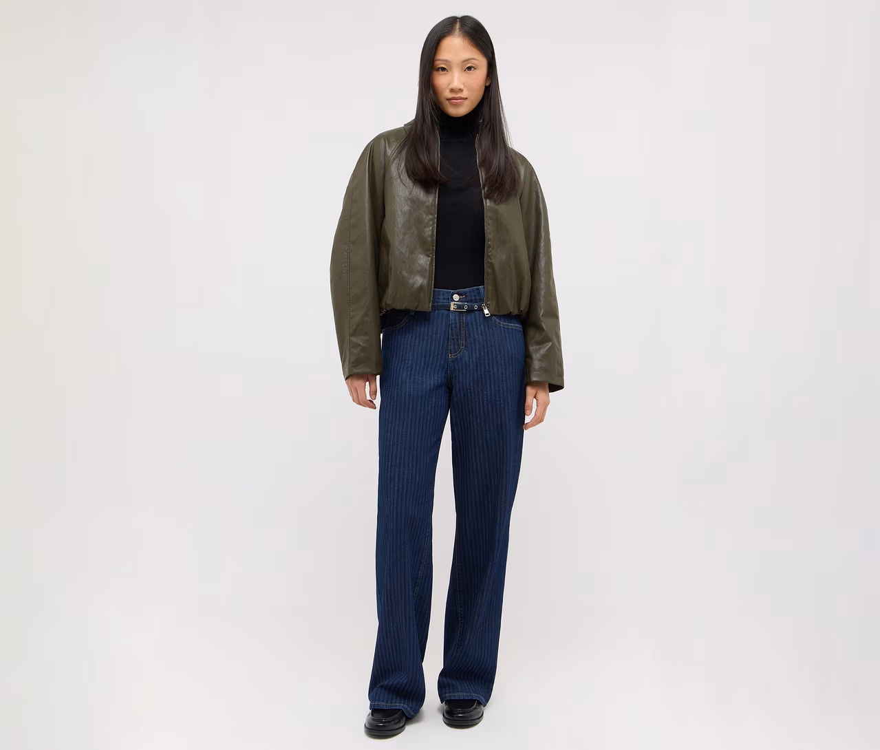 Femme debout portant une veste bomber vert foncé, un col roulé noir, un jean bleu foncé à fines rayures avec une boucle argentée et des chaussures noires.