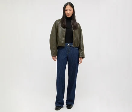 Femme debout portant une veste bomber vert foncé, un col roulé noir, un jean bleu foncé à fines rayures avec une boucle argentée et des chaussures noires.