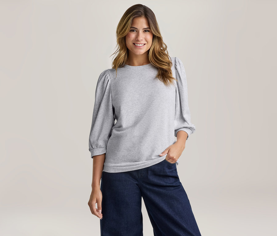 Une femme souriante aux longs cheveux ondulés porte un sweat-shirt gris clair à manches bouffantes et un pantalon large bleu foncé.