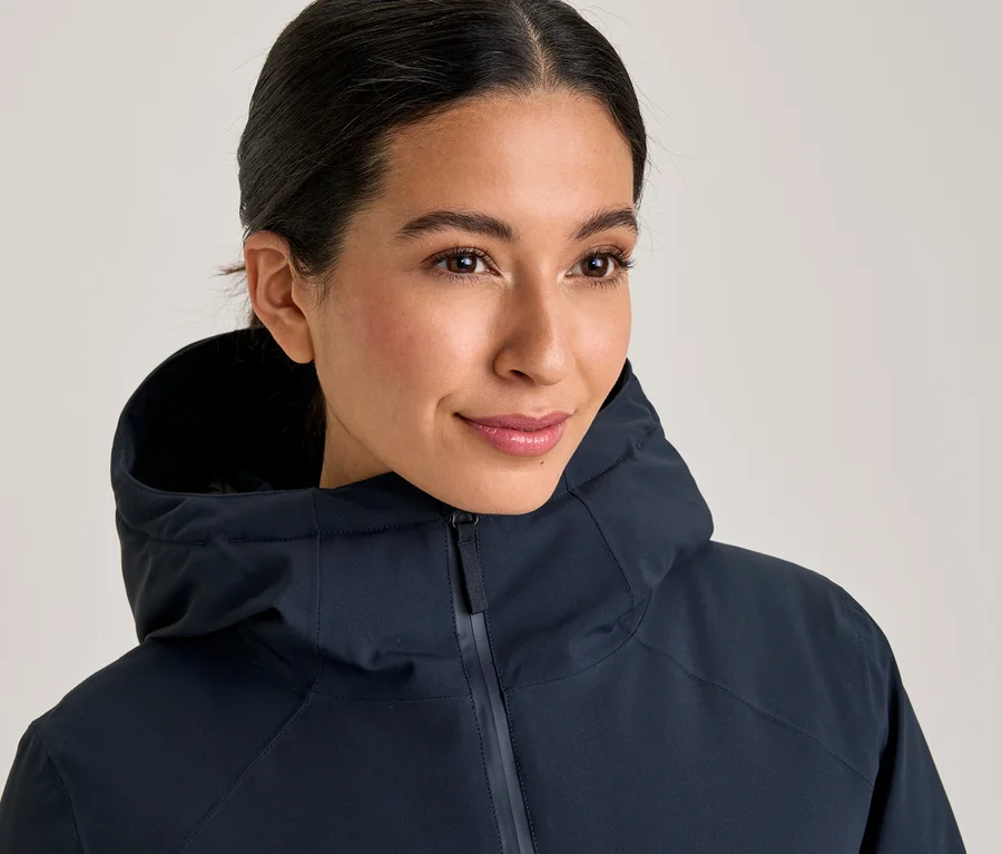 Une femme souriante aux cheveux foncés attachés porte une veste à capuche bleu foncé, zippée jusqu'au cou.