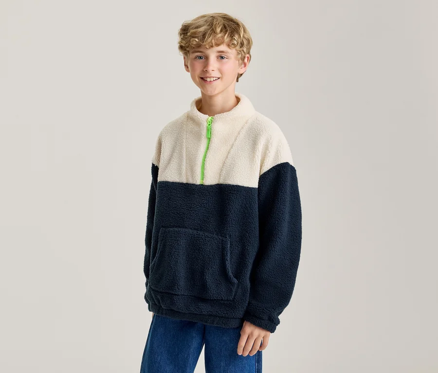 Garçon souriant portant un pull polaire bicolore crème et bleu marine avec une fermeture éclair vert vif et un jean bleu.
