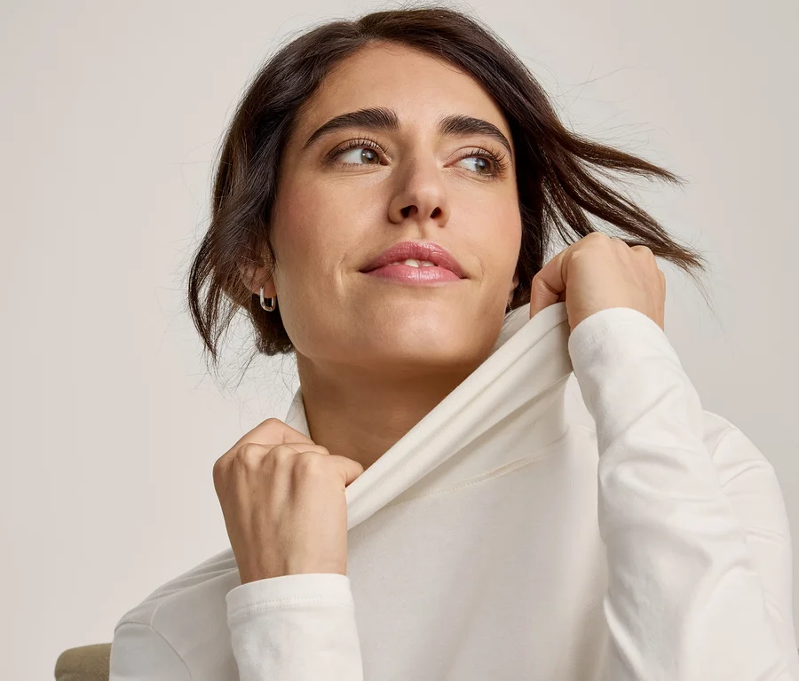 Femme aux cheveux foncés portant un haut blanc à manches longues, remontant le col et regardant vers la droite.