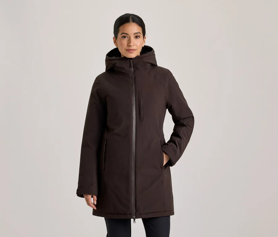 Femme aux cheveux foncés attachés en arrière, debout de face, portant une veste à capuche marron foncé avec les mains dans les poches.