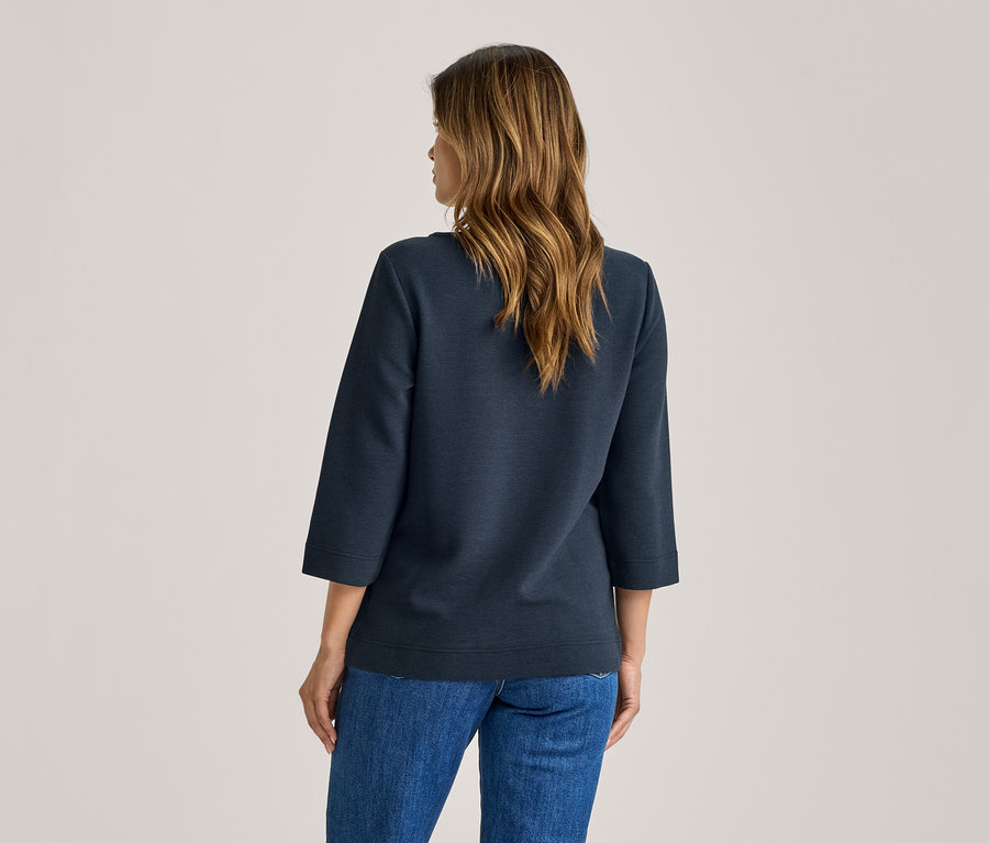 Femme de dos, portant un haut bleu foncé à manches trois-quarts et un jean bleu. Elle a de longs cheveux bruns ondulés.