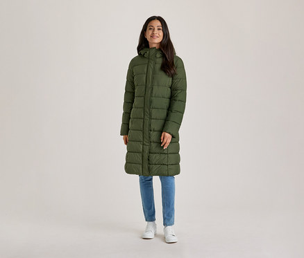 Une femme souriante aux cheveux foncés se tient de face, vêtue d'un long manteau matelassé vert olive à capuche, d'un jean bleu et de baskets blanches.