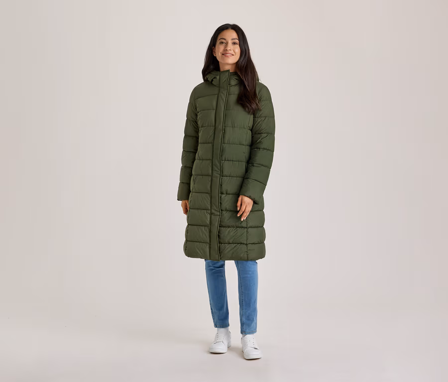 Une femme souriante aux cheveux foncés se tient de face, vêtue d'un long manteau matelassé vert olive à capuche, d'un jean bleu et de baskets blanches.