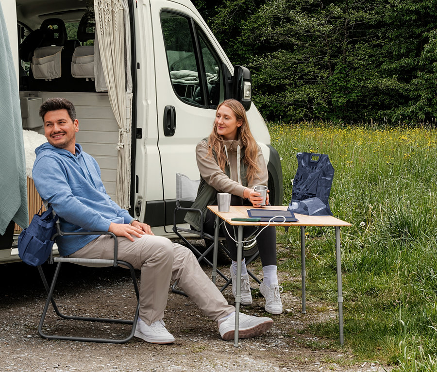 Un homme en sweat-shirt à capuche bleu clair et une femme en veste en sweat beige et gilet hybride kaki sont assis devant un camping-car. Un gilet pliable bleu foncé est posé sur une table.