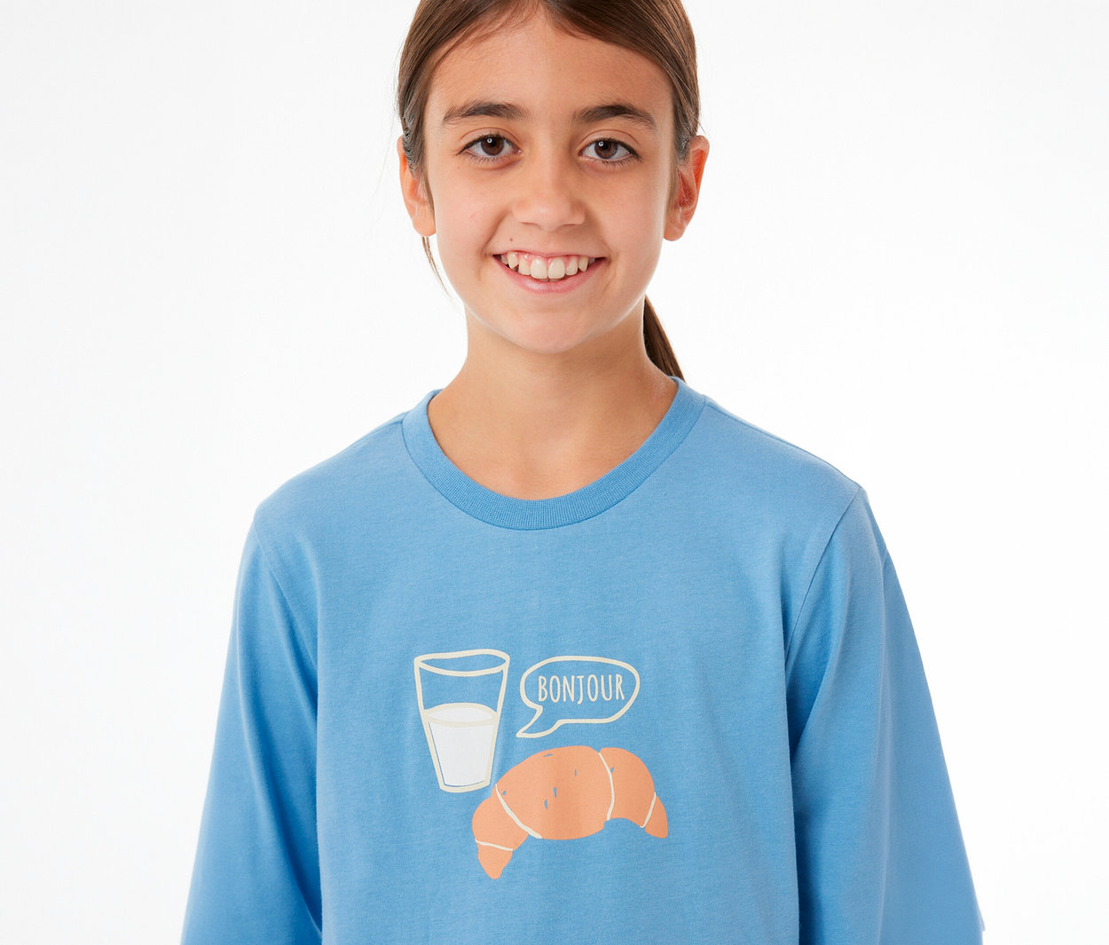 Jeune fille souriante portant un haut de pyjama bleu à manches longues, avec un motif de verre de lait, un croissant et une bulle de dialogue « BONJOUR ».
