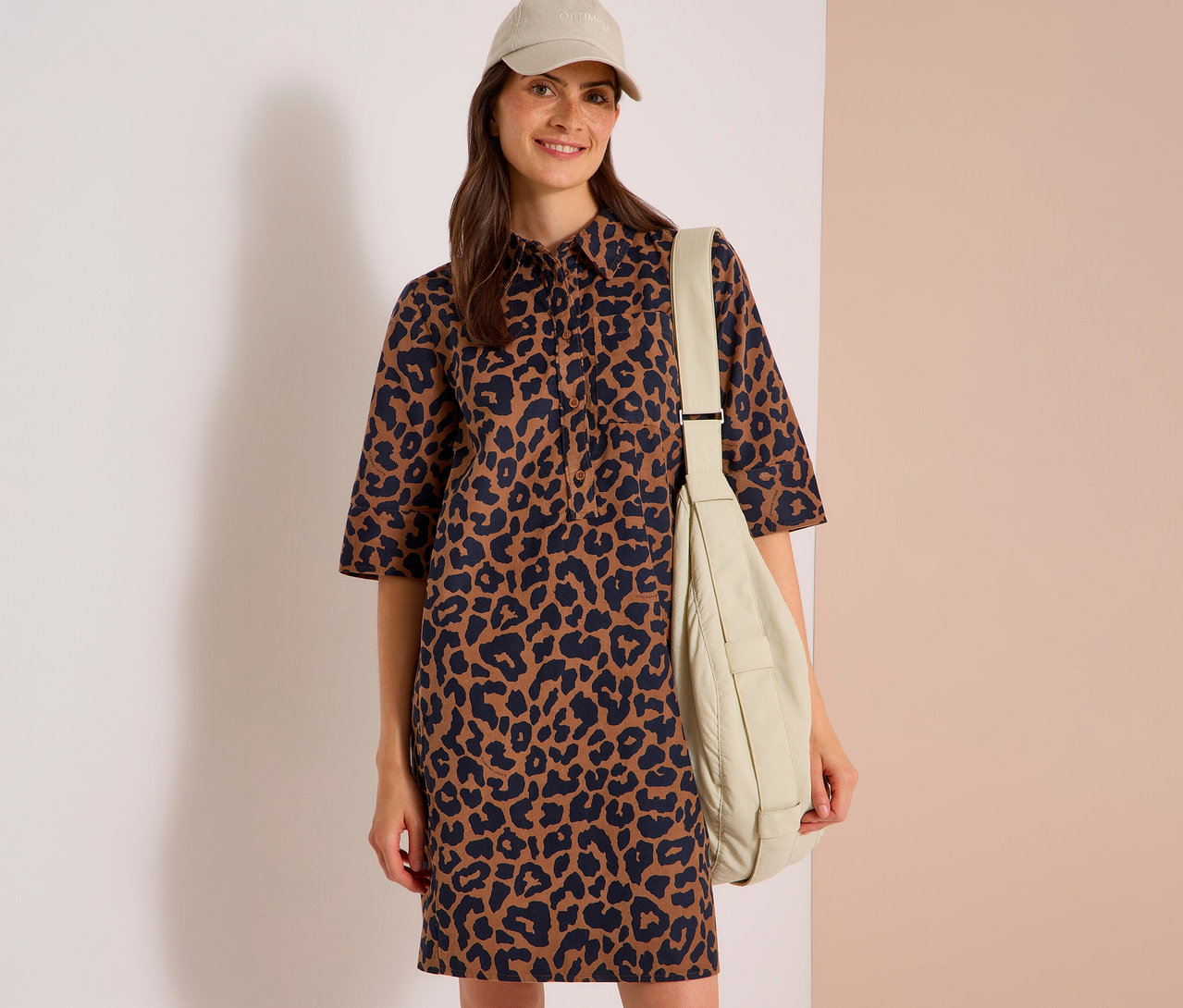Femme souriante portant une robe léopard marron et noire, une casquette de baseball beige et un sac à bandoulière beige.