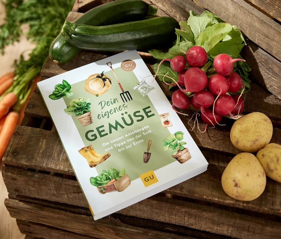 Le livre « Dein eigenes Gemüse » (tes propres légumes, en allemand) est posé sur une caisse en bois, entouré de carottes, de courgettes, de radis et de pommes de terre.