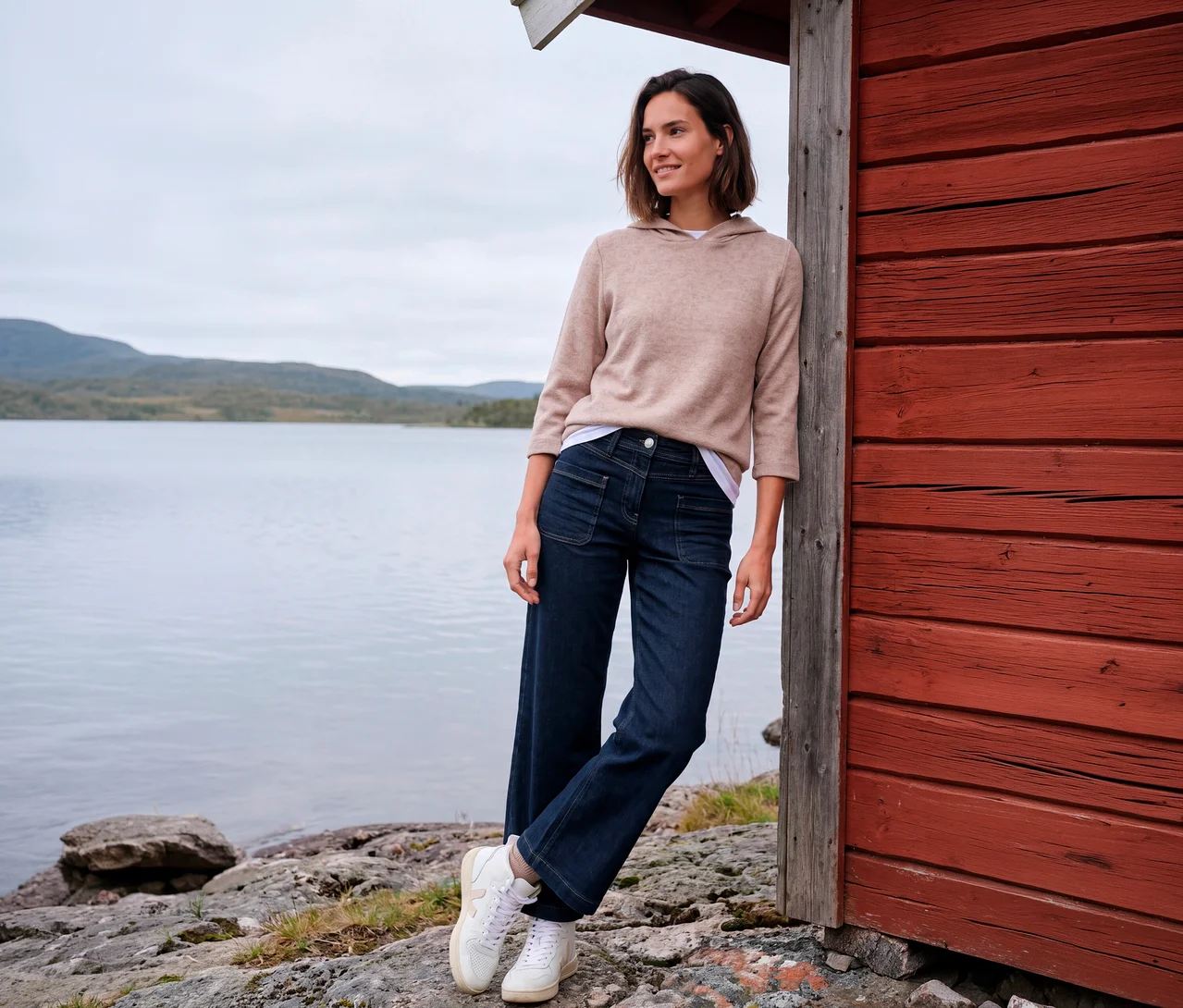 Une femme souriante en sweat à capuche beige, jean CECIL bleu foncé à jambe large et baskets blanches s'appuie sur une cabane rouge au bord d'un lac avec des montagnes.