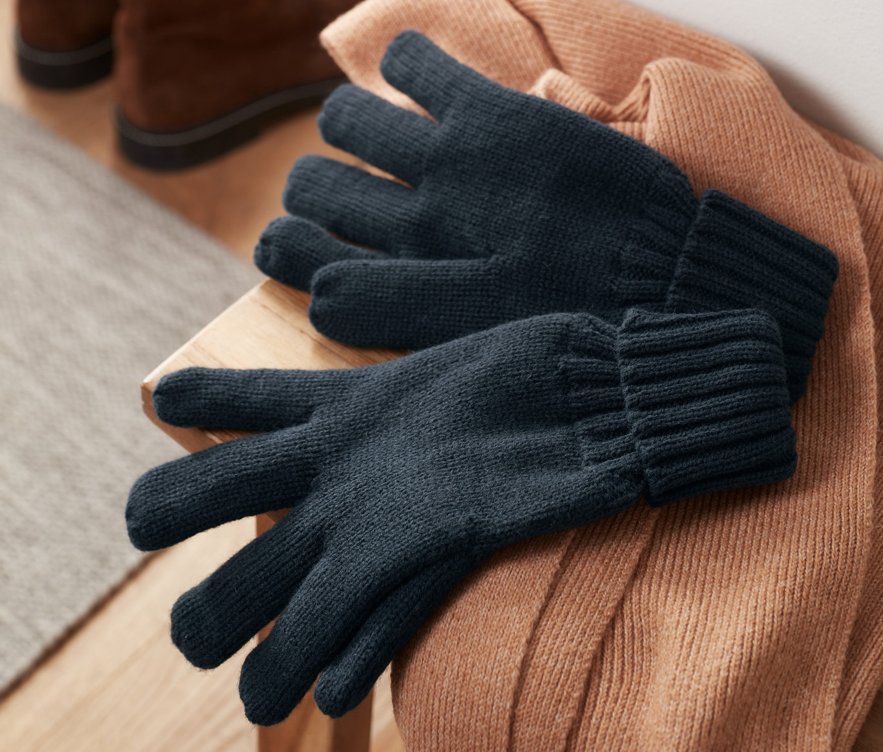 Des gants en maille posés sur un pull marron, avec des chaussures en arrière-plan.