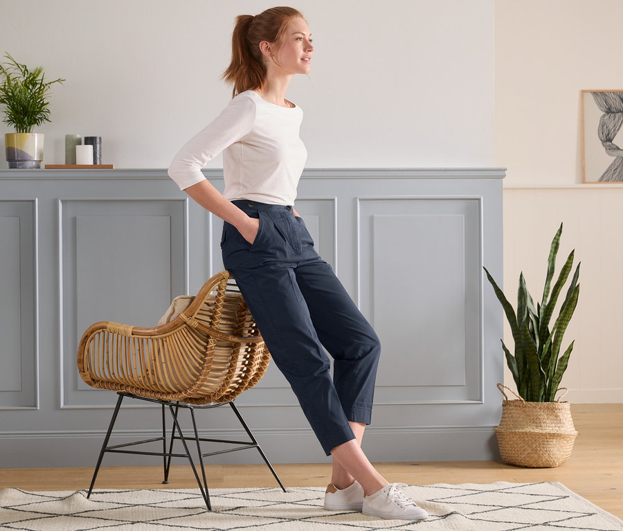 Une femme appuyée contre un fauteuil en rotin porte un chino moderne bleu foncé.