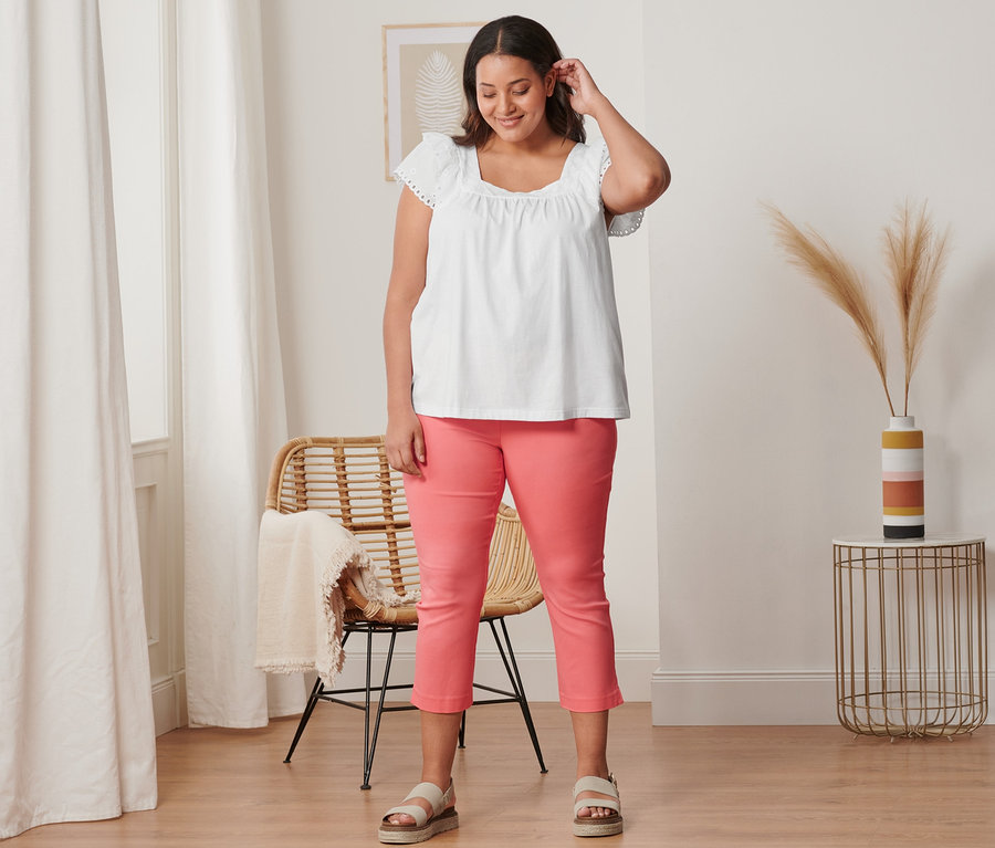 Une femme porte un pantalon stretch 7/8 corail, un t-shirt blanc à broderies et des sandales à plateforme en cuir.