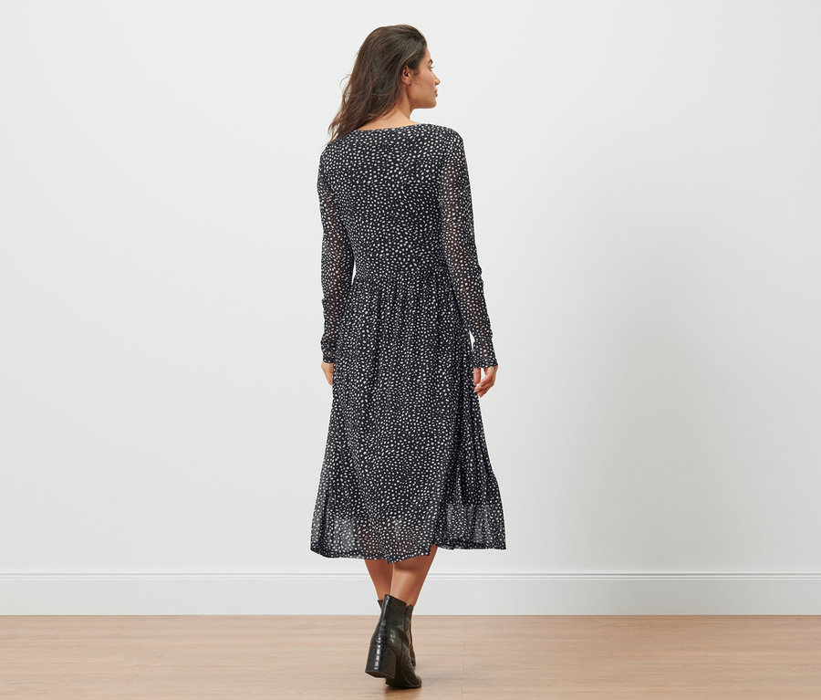 Une femme vue de dos marche sur le sol dans une robe en mesh à pois et des bottes noires.