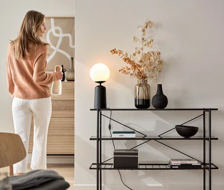 Femme tenant une bouteille à côté d'une console (largeur env. 120 cm) avec lampe de table noire avec globe en verre.