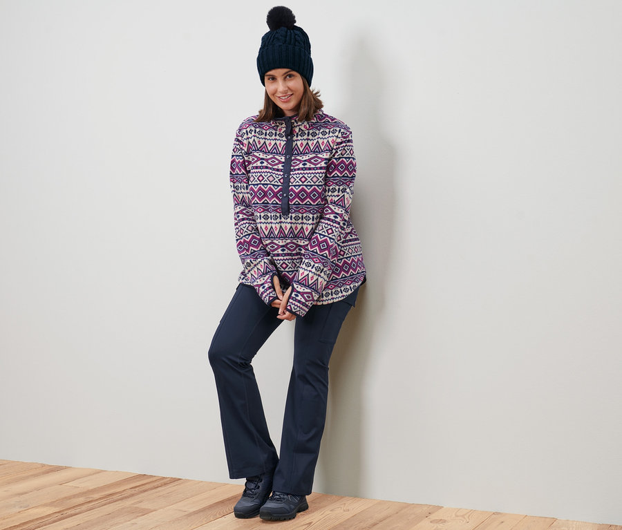 Une femme appuyée contre un mur porte un pull en polaire, un bonnet en maille et un pantalon jazz thermique.