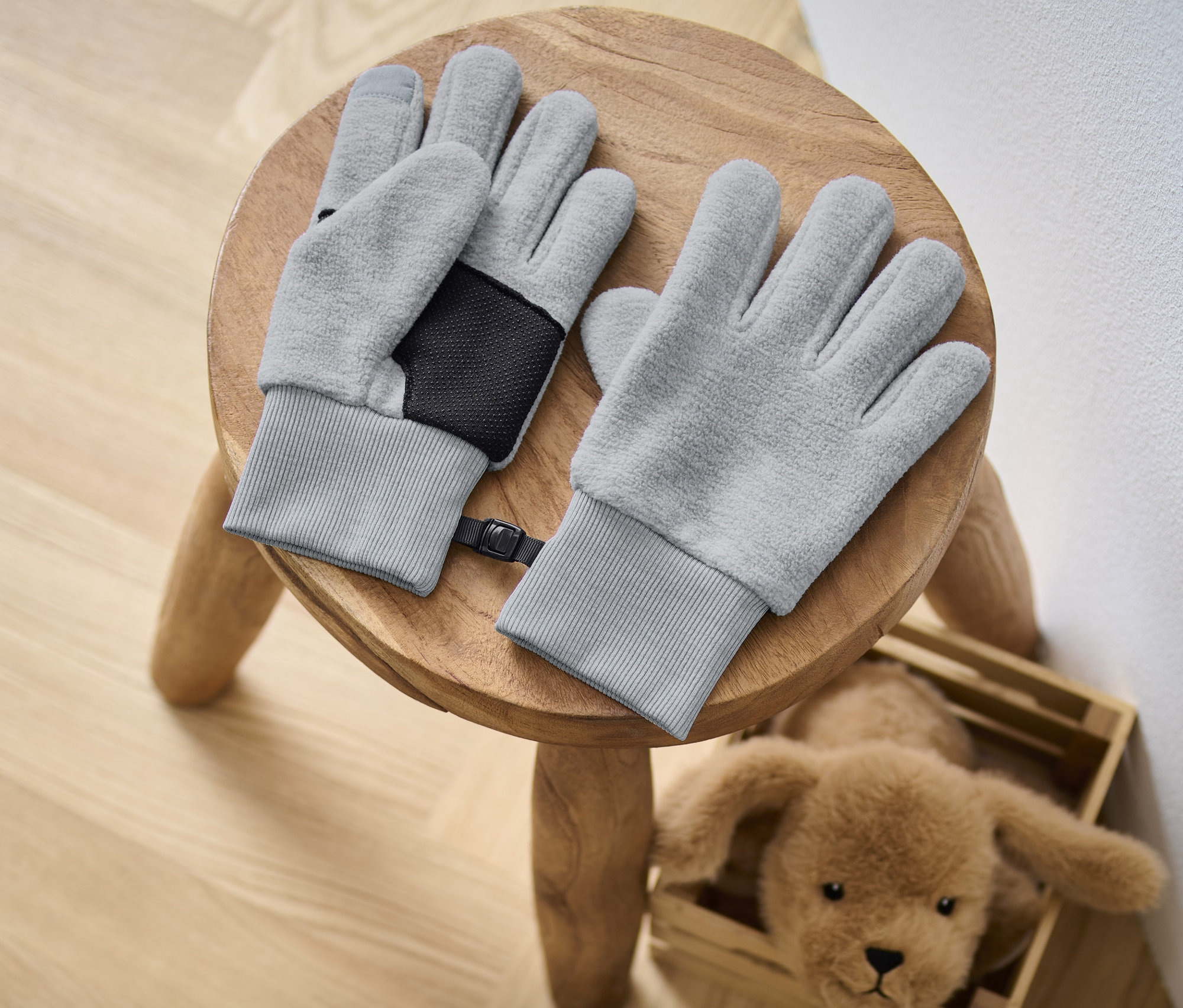 Deux gants en polaire pour enfant sont posés sur un tabouret en bois à côté d'un chien en peluche.