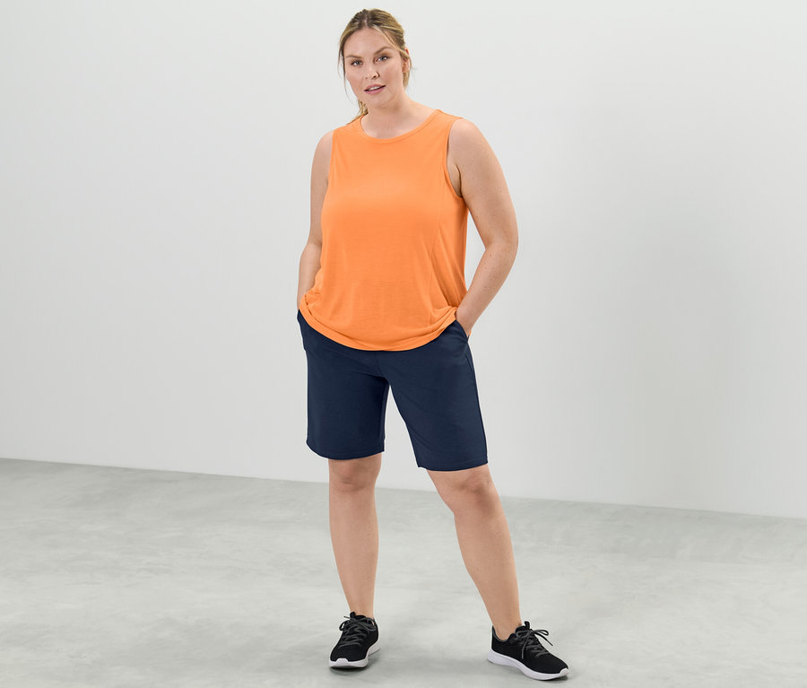 Une femme se tient debout, les mains dans les poches de son short, portant un top de sport pêche d'un ensemble deux pièces et un short bleu marine.