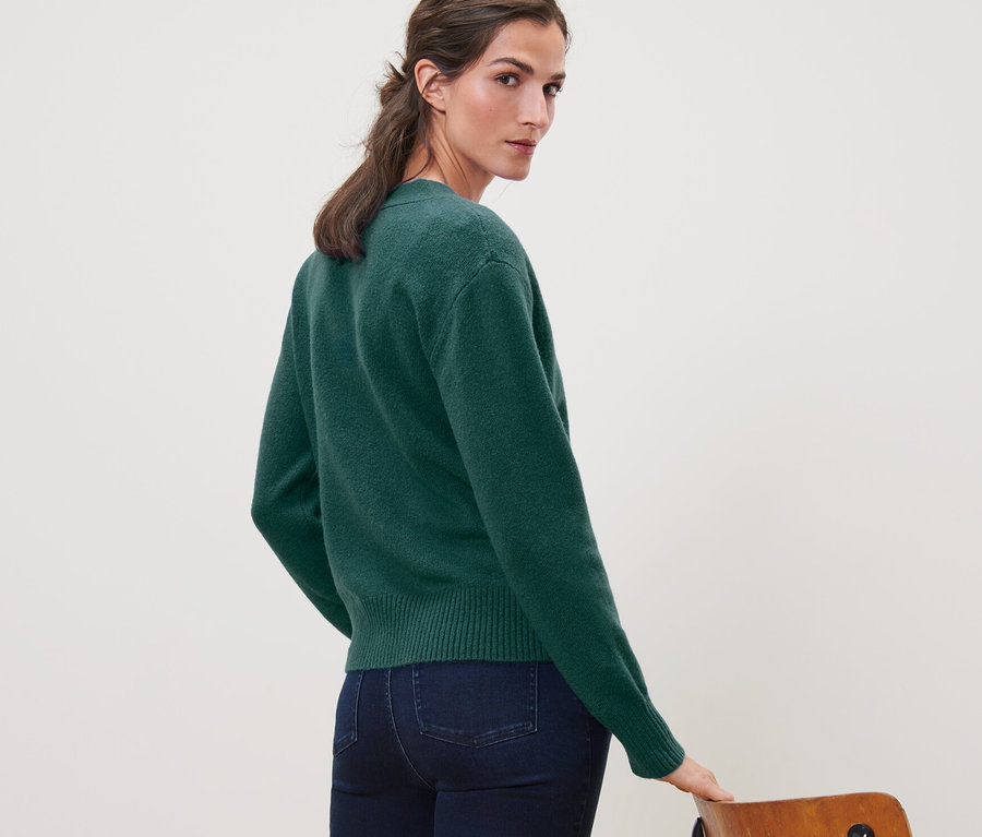 Une femme regarde par-dessus son épaule et porte une jaquette en maille avec de la laine vert foncé et un jean.