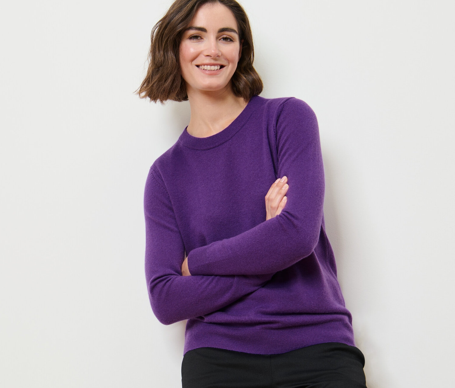 Une femme pose, les bras croisés, portant un pull violet en maille fine avec du cachemire et un pantalon noir.