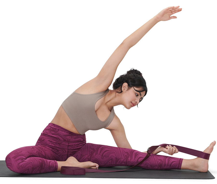 Une femme est assise sur un tapis de yoga et utilise une ceinture de yoga 2 en 1 violette pour étirer sa jambe.
