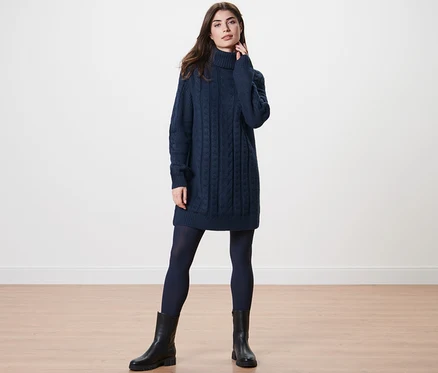 Femme posant en pull long en maille avec torsades, collants sombres et bottes noires.