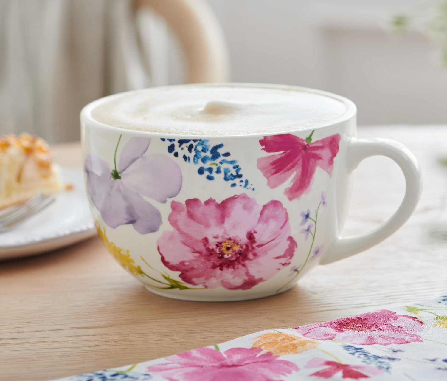 Sur une table, un bol à café au lait à motifs floraux et une part de gâteau avec de la crème chantilly.