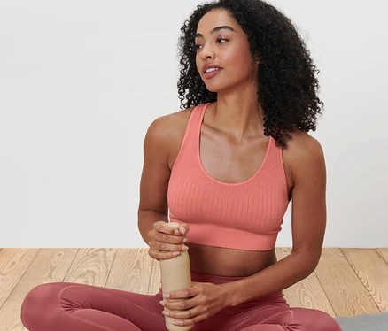 Une femme est assise les jambes croisées sur un tapis de yoga et tient une bouteille d'eau. Elle porte une brassière de sport sans coutures vieux rose.